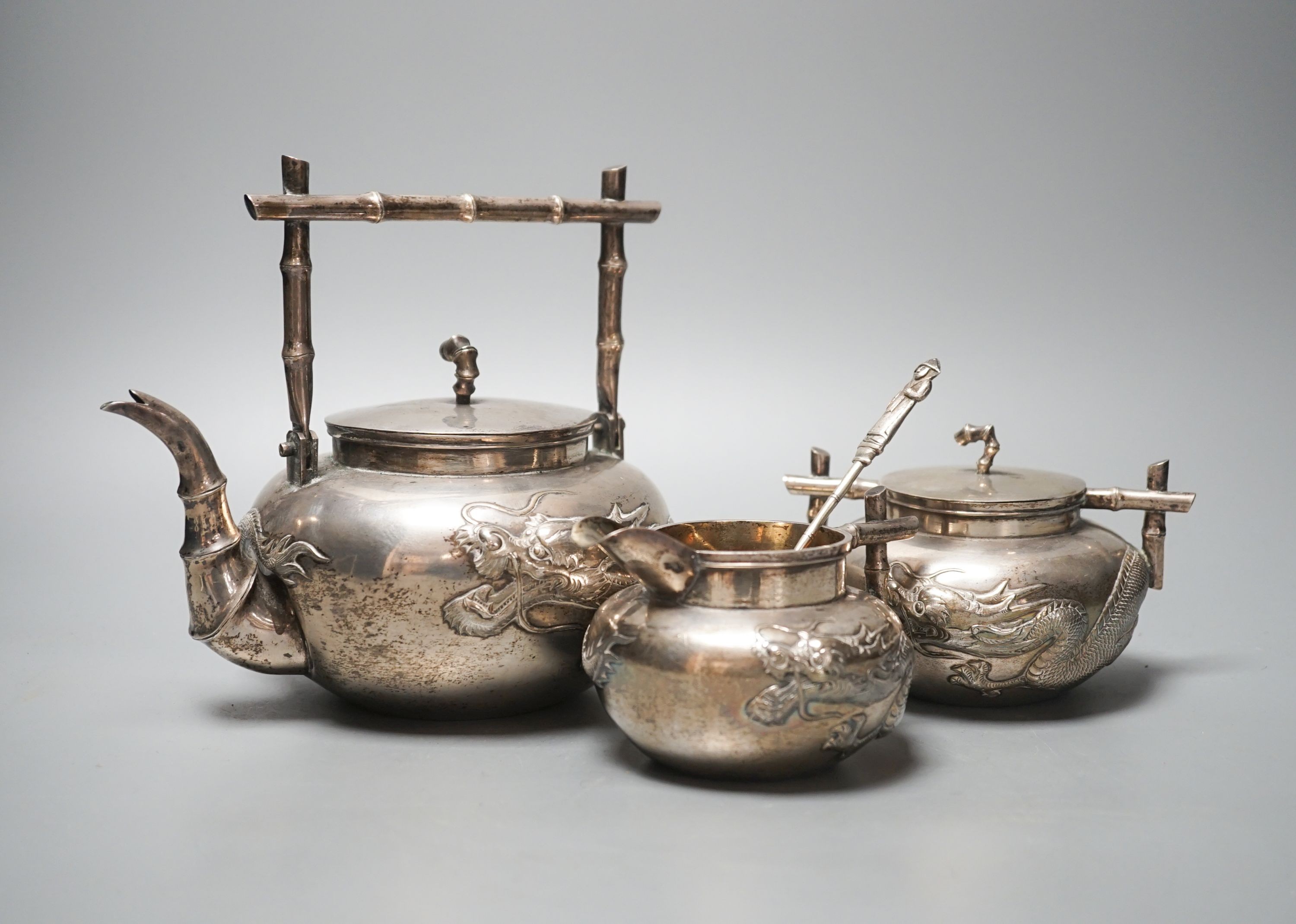 A Chinese white metal three piece tea set, with embossed dragon decoration and faux bamboo handles, teapot height, 17.8cm, gross 28.5oz (a.f.) and a similar spoon.